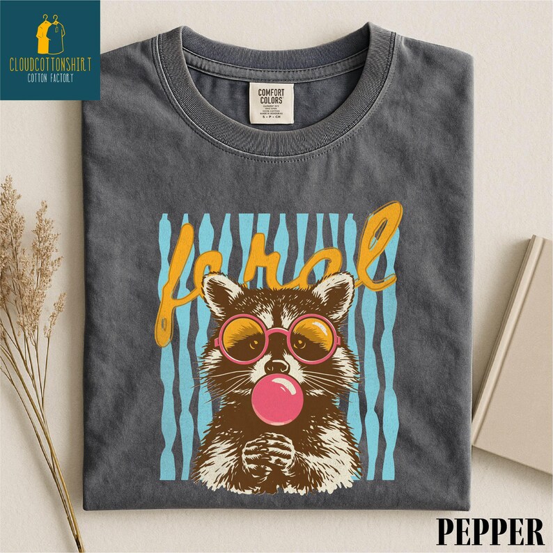 A gray t-shirt with a graphic design featuring a raccoon wearing pink bubblegum and sunglasses, along with the word "Pepper" written above it.