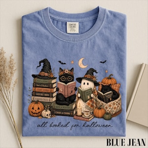 A blue t-shirt with a Halloween-themed illustration of a witch, cat, ghost, pumpkin, and books, along with the text "all booked for Halloween" and the brand name "BLUE JEAN".