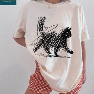 May include: Ivory-colored t-shirt featuring a black scribble cat design. The shirt has short sleeves and a crew neck. The text "CLOUDCOTTONSHIRT COTTON FACTORY" is on the upper left. The word "IVORY" is at the bottom right.