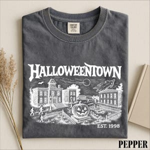 A gray t-shirt with a Halloween-themed graphic design, including a spooky town scene, a jack-o-lantern, and a cemetery.