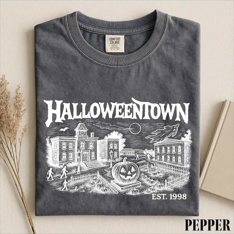 A gray t-shirt with a Halloween-themed graphic design, including a spooky town scene, a jack-o-lantern, and a cemetery.