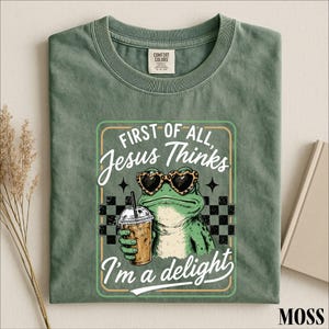 May include: A sage green crew neck t-shirt featuring a graphic of a green frog wearing leopard print heart-shaped sunglasses and holding an iced coffee. The text reads: First of all, Jesus Thinks I'm a delight. The design includes a black checkerboard.