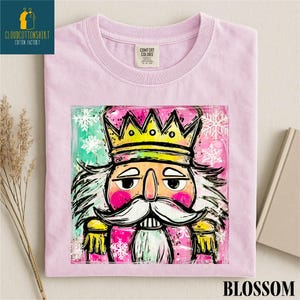 Może przedstawiać: Jasnor&oacute;żowa koszulka z grafiką Kr&oacute;la Dziadka do Orzech&oacute;w w złotej koronie. Grafika ma r&oacute;żowe, zielone i białe akcenty. Słowo "BLOSSOM" jest wydrukowane na dole.
