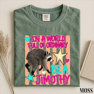 A green t-shirt with a colorful graphic design featuring a raccoon and text that reads "In a world full of ordinary, be a jimothy".