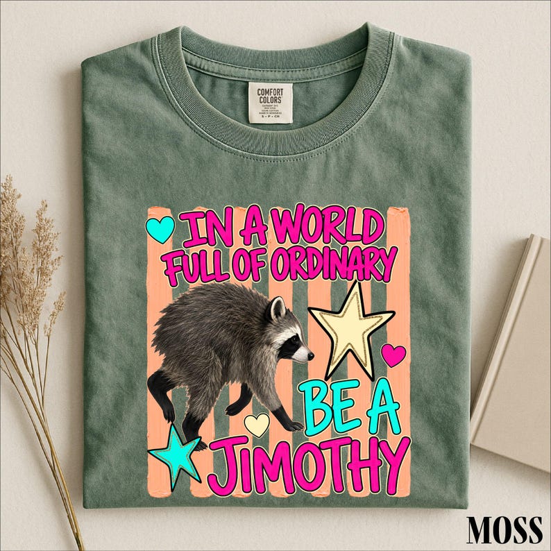 A green t-shirt with a colorful graphic design featuring a raccoon and text that reads "In a world full of ordinary, be a jimothy".