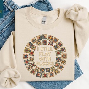 May include: Sand-colored sweatshirt with the text "I STILL PLAY WITH BLOCKS" in the center, surrounded by a circular design of colorful quilt blocks. The sweatshirt is displayed on a pair of blue jeans.