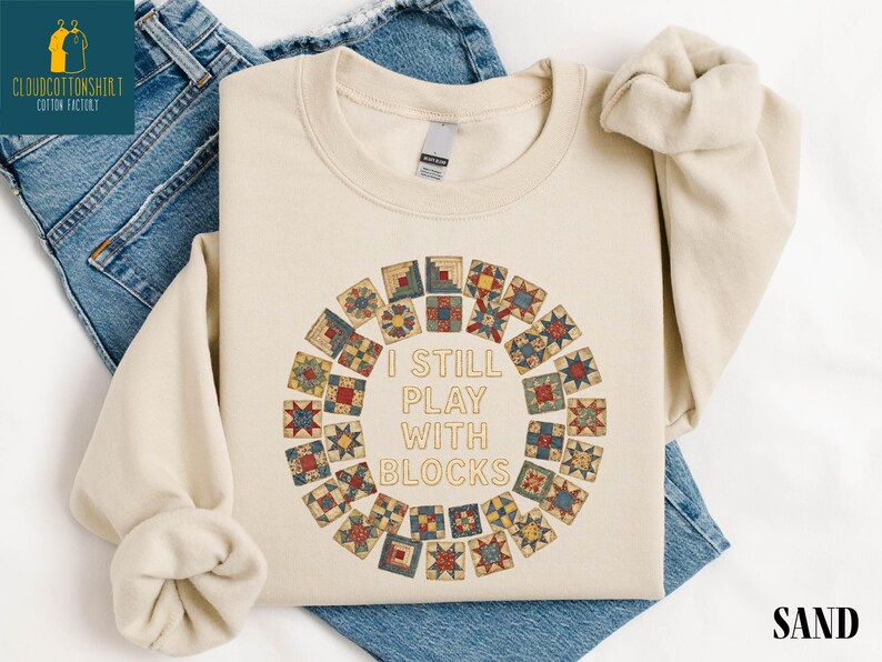 May include: Sand-colored sweatshirt with the text "I STILL PLAY WITH BLOCKS" in the center, surrounded by a circular design of colorful quilt blocks. The sweatshirt is displayed on a pair of blue jeans.