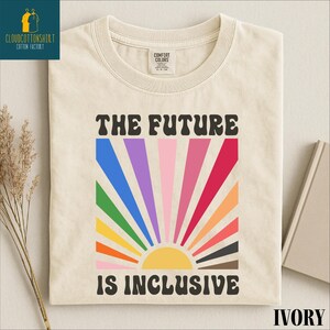 May include: Ivory-colored t-shirt with the text "THE FUTURE IS INCLUSIVE" in black. The design features a colorful sunburst with rainbow-colored rays. The shirt has a crew neck and short sleeves.