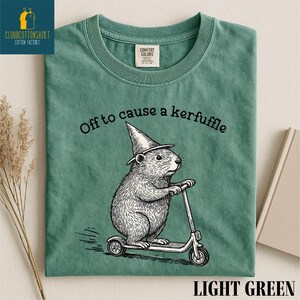 A green t-shirt with a cartoon image of a mouse riding a scooter, wearing a pointy hat, and the text "Off to cause a kerfuffle" printed on it.