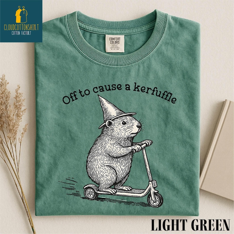 A green t-shirt with a cartoon image of a mouse riding a scooter, wearing a pointy hat, and the text "Off to cause a kerfuffle" printed on it.