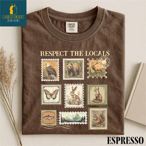 May include: Espresso-colored t-shirt with the phrase "Respect the Locals" above a grid of vintage-style postage stamps featuring wildlife like a bear, eagle, and fox. The shirt has a crew neck and a Comfort Colors tag.