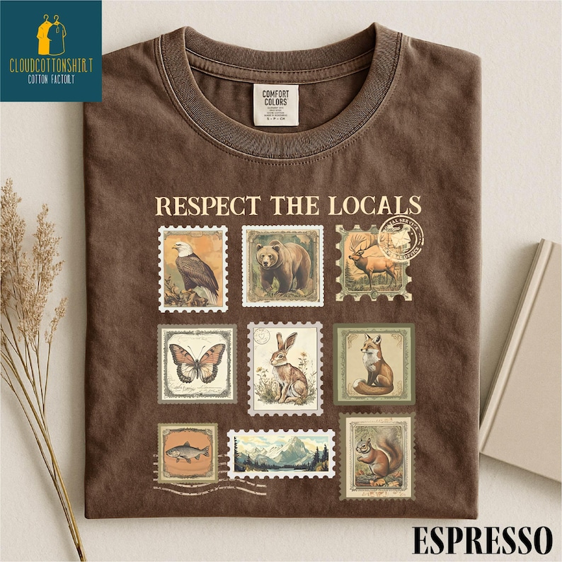 May include: Espresso-colored t-shirt with the phrase "Respect the Locals" above a grid of vintage-style postage stamps featuring wildlife like a bear, eagle, and fox. The shirt has a crew neck and a Comfort Colors tag.