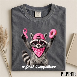 May include: A charcoal gray t-shirt featuring a graphic of a raccoon wearing a pink baseball cap, heart-shaped sunglasses, and a pink bandana with ribbon patterns. The raccoon holds a pink ribbon and a donut, with the text feral & supportive.