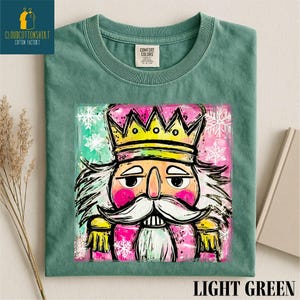 Może przedstawiać: Jasnozielona koszulka z kolorową ilustracją dziadka do orzech&oacute;w ze złotą koroną, r&oacute;żowymi policzkami i białymi wąsami. Koszulka ma napis "LIGHT GREEN" na dole. Koszulka jest marki Comfort Colors.