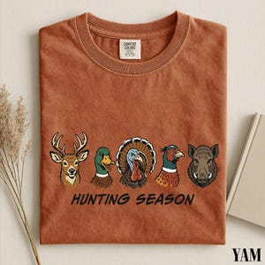 a brown t-shirt with a graphic design of various bird species, including a turkey, a duck, a goose, and a boar, along with the text "HUNTING SEASON".