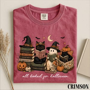 A pink t-shirt with a Halloween-themed illustration of cats reading books, surrounded by pumpkins, gourds, and bats.