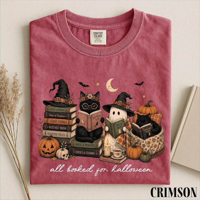 A pink t-shirt with a Halloween-themed illustration of cats reading books, surrounded by pumpkins, gourds, and bats.