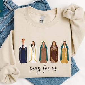 May include: Sand-colored sweatshirt featuring an illustration of five religious figures in various robes and crowns. The phrase "pray for us" is written in cursive below the figures. The sweatshirt is displayed on a pair of blue jeans.
