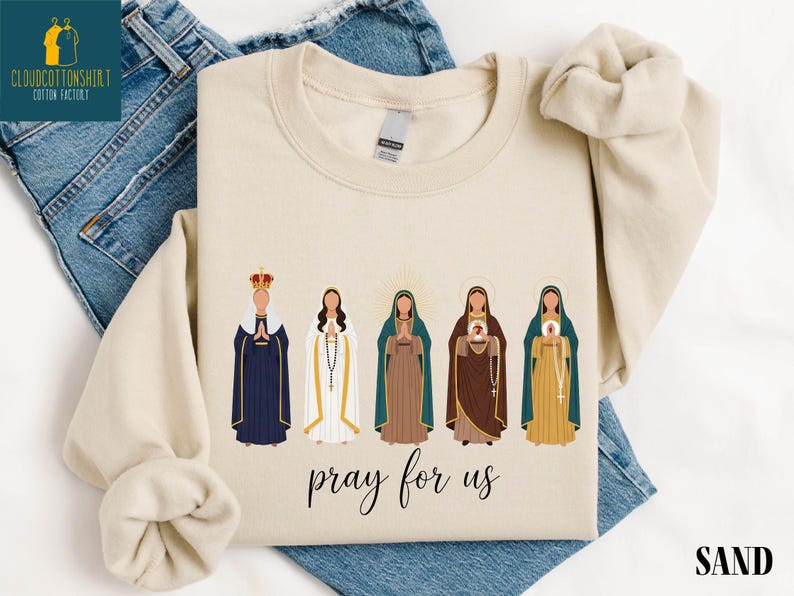 May include: Sand-colored sweatshirt featuring an illustration of five religious figures in various robes and crowns. The phrase "pray for us" is written in cursive below the figures. The sweatshirt is displayed on a pair of blue jeans.