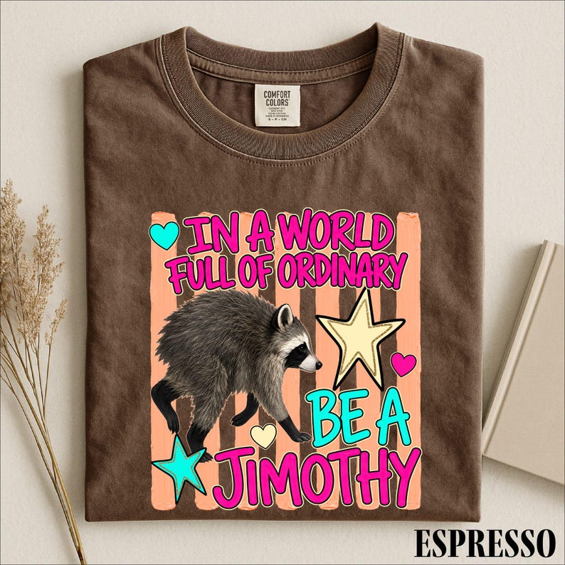 A brown t-shirt with a colorful graphic design featuring a raccoon and the text "In a world full of ordinary, be a jimothy".
