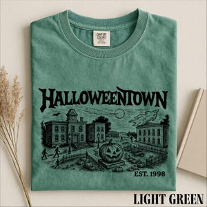 A green t-shirt with a Halloween-themed graphic design, including a pumpkin patch, a castle, and a ghost.