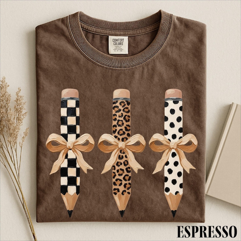 a brown t-shirt with three pencils, each with a unique design and a bow, arranged in a row.