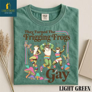Pu&ograve; includere: T-shirt verde chiaro con la scritta "They Turned The Frigging Frogs Gay" e una grafica di tre rane che ballano. Le rane indossano abiti colorati e sono circondate da farfalle e funghi.