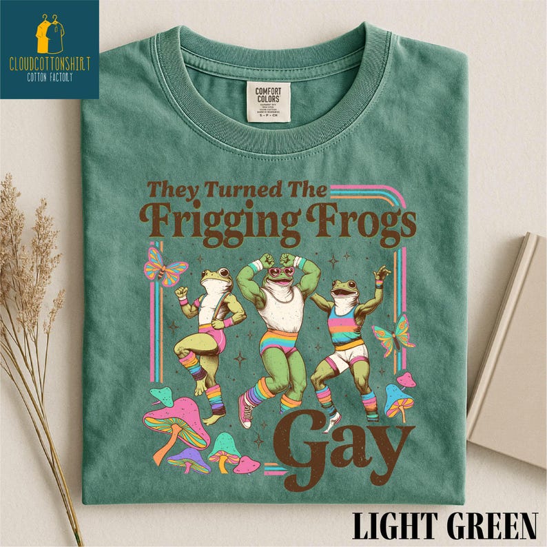 Pu&ograve; includere: T-shirt verde chiaro con la scritta "They Turned The Frigging Frogs Gay" e una grafica di tre rane che ballano. Le rane indossano abiti colorati e sono circondate da farfalle e funghi.