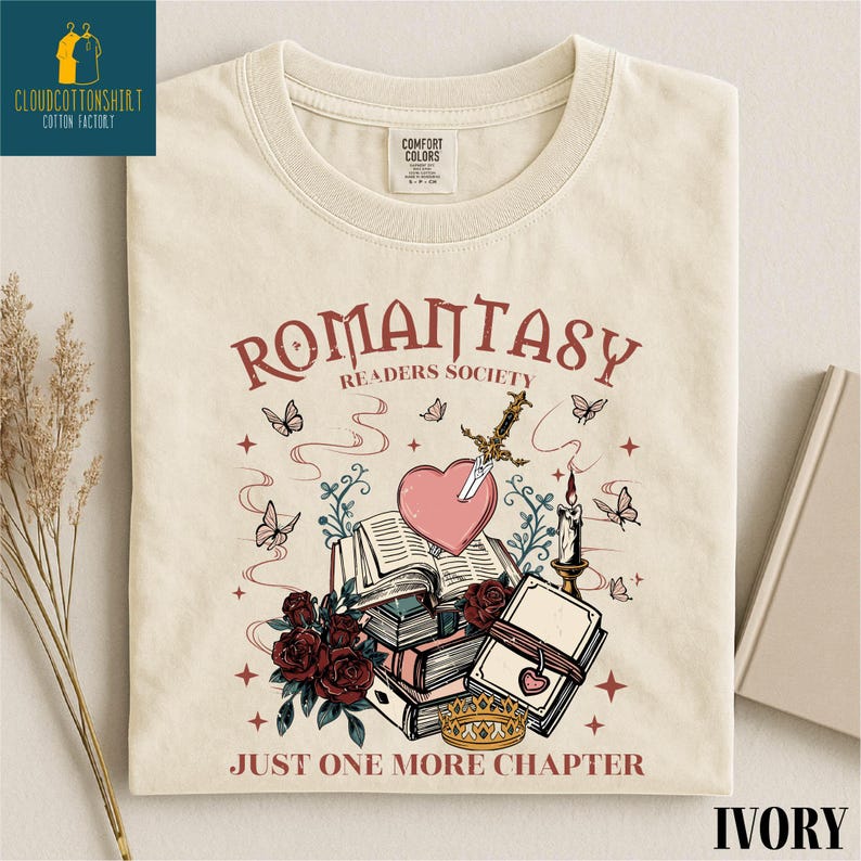 May include: Ivory-colored t-shirt with a graphic design featuring the text "ROMANTASY READERS SOCIETY" and "JUST ONE MORE CHAPTER." The design includes a heart pierced by a sword, books, roses, butterflies, and a crown.