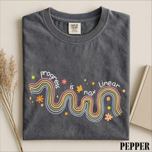 May include: A charcoal gray crew neck t-shirt featuring a graphic of a wavy, multi-colored rainbow line with the text "progress is not linear" written in white. The design includes small orange and yellow flower icons and star shapes.