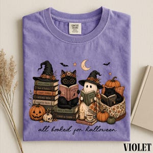 A purple t-shirt with a Halloween-themed illustration of a witch, cat, ghost, pumpkin, and books, along with the text "all read for Halloween".