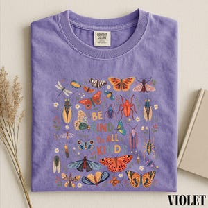 A purple t-shirt with a colorful graphic design of various insects and butterflies, along with the text "Be kind to all kids" printed on it.