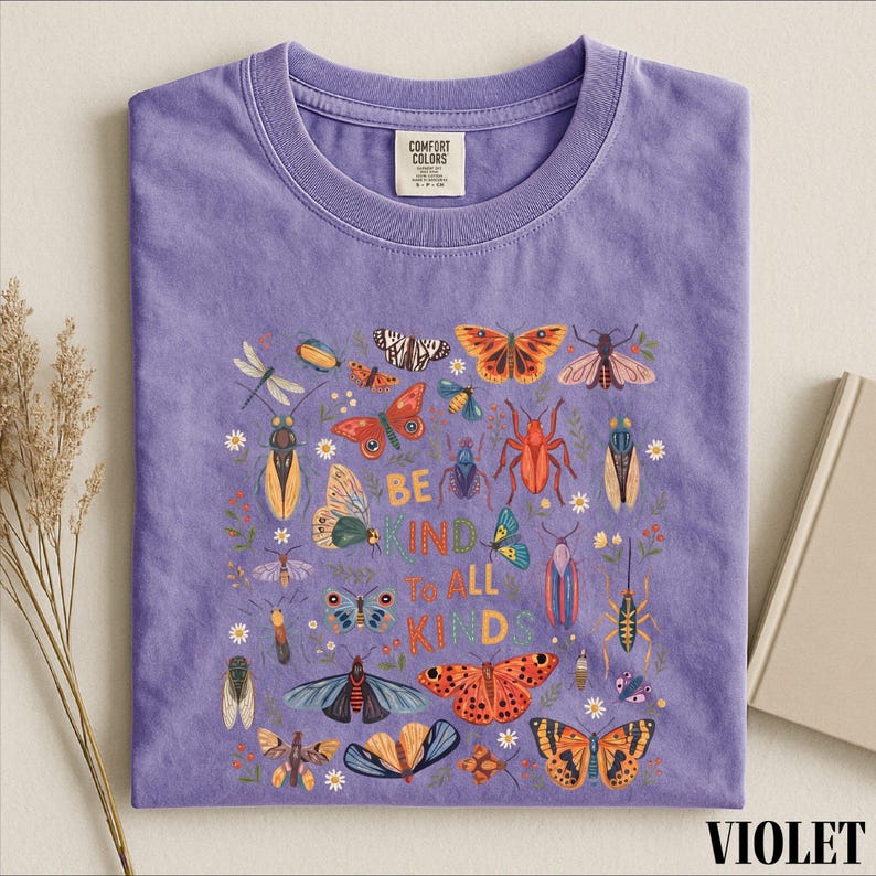 A purple t-shirt with a colorful graphic design of various insects and butterflies, along with the text "Be kind to all kids" printed on it.