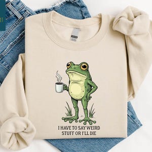 A beige t-shirt with a cartoon frog holding a coffee cup and the text "I HAVE TO SAY WEIRD STUFF OR ILL DIE" printed on it.
