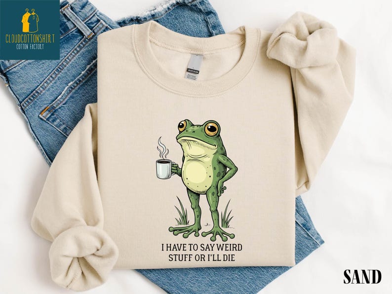 A beige t-shirt with a cartoon frog holding a coffee cup and the text "I HAVE TO SAY WEIRD STUFF OR ILL DIE" printed on it.