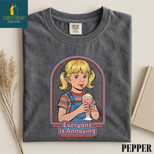a gray t-shirt with a graphic design depicting a young girl with blonde hair and a straw, holding a pink drink. The text below the graphic reads "Everyone is Annoying".