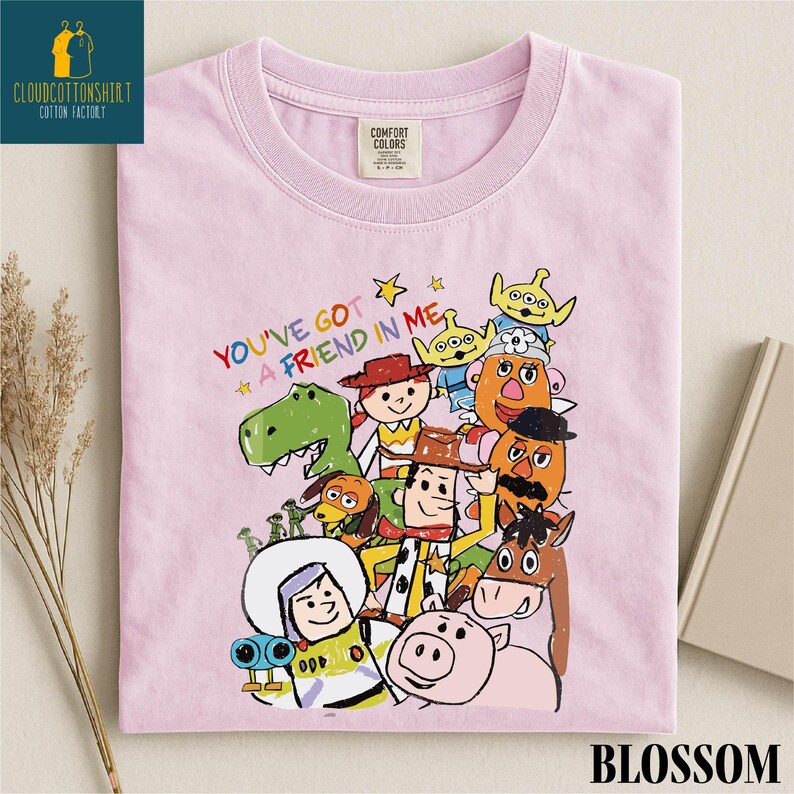 K&ouml;nnte beinhalten: Hellrosa T-Shirt mit einer bunten Cartoon-Illustration von Charakteren aus "Toy Story". Der Text "YOU'VE GOT A FRIEND IN ME" steht &uuml;ber den Figuren. Das T-Shirt hat einen Rundhalsausschnitt und kurze &Auml;rmel.