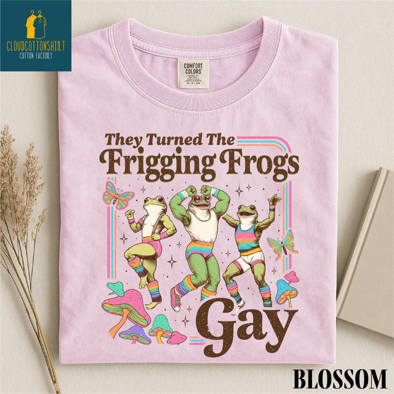 Pu&ograve; includere: T-shirt rosa chiaro con la scritta "They Turned The Frigging Frogs Gay" in marrone. La maglietta presenta un disegno di tre rane in abbigliamento sportivo colorato, con farfalle e funghi. La maglietta ha un'etichetta Comfort Colors.
