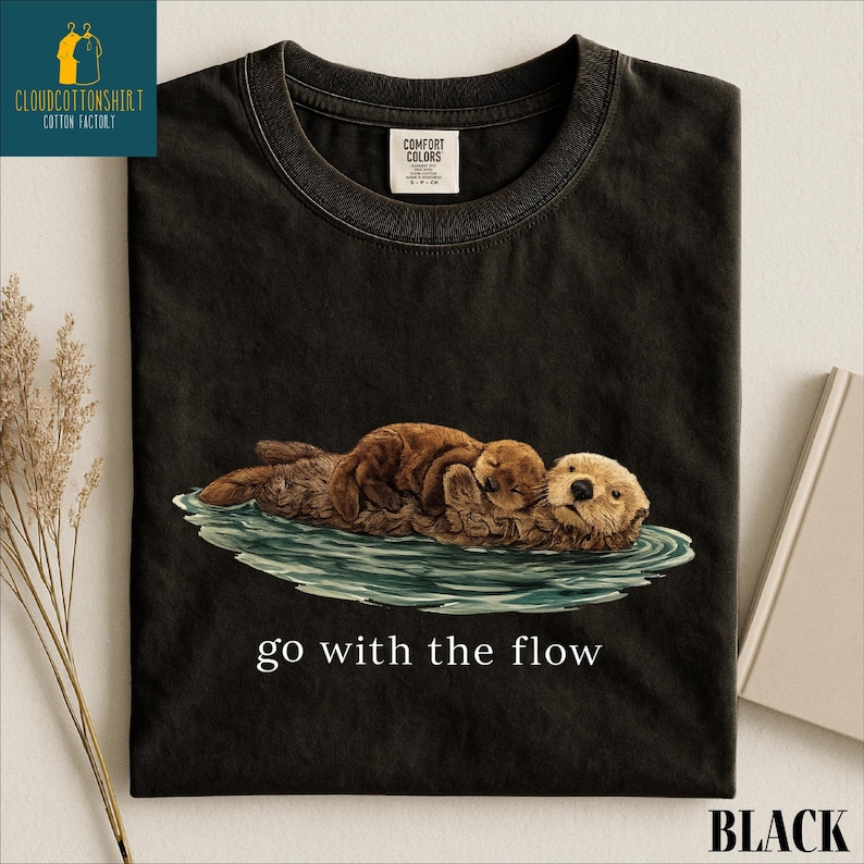 A black t-shirt with a printed image of two otters swimming together in a body of water, accompanied by the text "go with the flow".