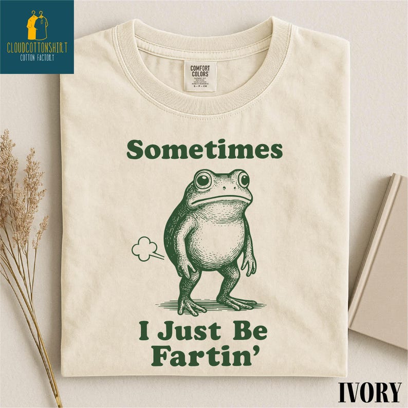 May include: Ivory-colored t-shirt with a green frog illustration and the text "Sometimes I Just Be Fartin'". The shirt features a crew neck and a Comfort Colors tag. The design is centered on the front of the shirt.