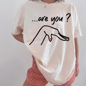 May include: An ivory short-sleeved t-shirt featuring the black text "...are you ?" above a black line-art illustration of a hand with red and multi-colored fingernails. The shirt is paired with red and white gingham patterned shorts.