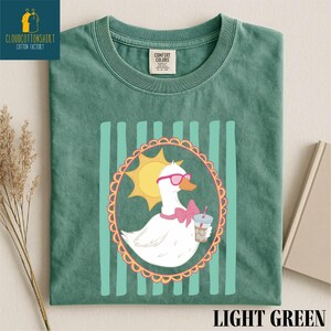 A green t-shirt with a cartoon image of a duck wearing sunglasses and holding a drink, surrounded by a decorative border.
