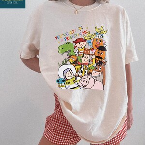 K&ouml;nnte beinhalten: Cremefarbenes T-Shirt mit einem farbenfrohen Grafikdesign von "Toy Story"-Charakteren und dem Text "You've Got a Friend in Me". Das Shirt wird mit rot-wei&szlig; karierten Shorts getragen. Das Wort "IVORY" ist unten aufgedruckt.