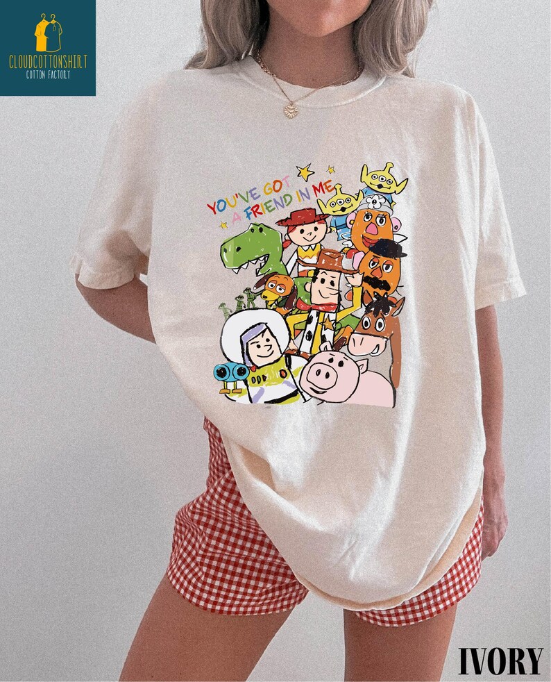 K&ouml;nnte beinhalten: Cremefarbenes T-Shirt mit einem farbenfrohen Grafikdesign von "Toy Story"-Charakteren und dem Text "You've Got a Friend in Me". Das Shirt wird mit rot-wei&szlig; karierten Shorts getragen. Das Wort "IVORY" ist unten aufgedruckt.