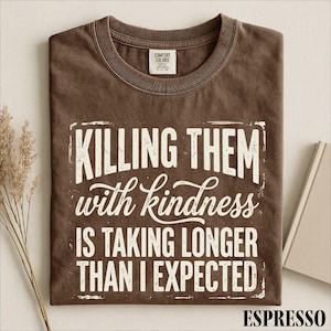 May include: A brown short-sleeved t-shirt with a crew neckline and a Comfort Colors label. The front features white distressed-style text that reads: KILLING THEM with kindness IS TAKING LONGER THAN I EXPECTED, enclosed in a rectangular border.