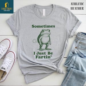 May include: Athletic heather gray t-shirt with a green illustration of a frog and the text "Sometimes I Just Be Fartin'". The shirt is paired with blue jeans and white sneakers.