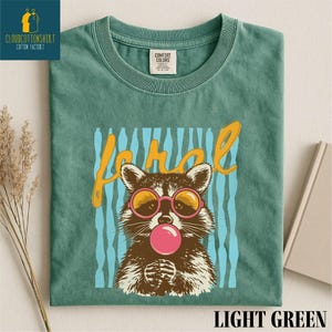 A green t-shirt with a graphic design of a raccoon wearing sunglasses and blowing a bubble gum bubble.