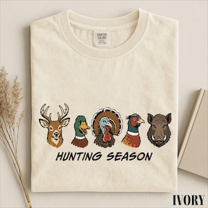 A beige t-shirt with a graphic design featuring illustrations of various hunting animals, including a deer, a turkey, a pheasant, a boar, and a duck, along with the text "HUNTING SEASON".