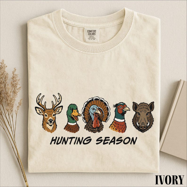 A beige t-shirt with a graphic design featuring illustrations of various hunting animals, including a deer, a turkey, a pheasant, a boar, and a duck, along with the text "HUNTING SEASON".