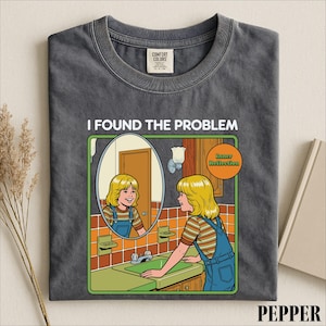 May include: A charcoal gray t-shirt featuring a graphic illustration of a person with blonde hair wearing blue overalls and a striped shirt, looking into a mirror. The text reads I FOUND THE PROBLEM and Inner Reflection in a circular orange badge.